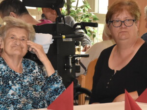 Zwei Frauen - eine Seniorin und ihre Angehörige - sitzen nebeneinander an einem Tisch. Im Hintergrund sind weitere Gäste in einem Gemeinschaftsraum zu sehen.