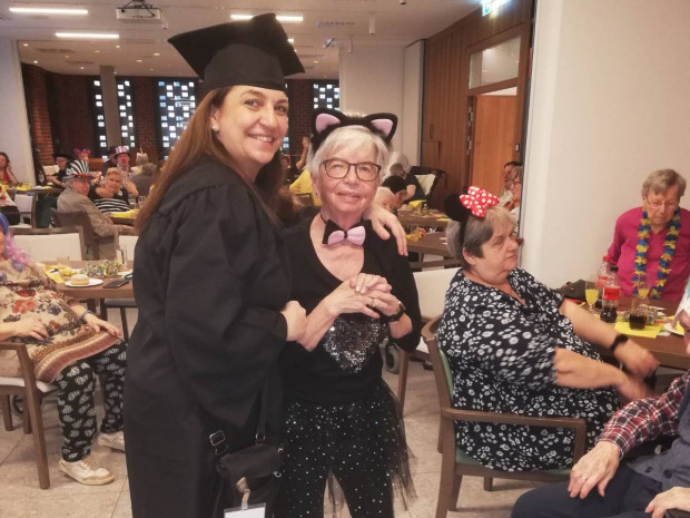 Eine als Absolventin verkleidete Frau hält die Hände einer älteren Person mit Katzenohren und Glitzerkostüm. Beide lächeln. Im Hintergrund sitzen weitere verkleidete Bewohner:innen an festlich dekorierten Tischen und genießen das bunte Faschingstreiben. 