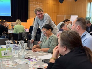 Mehrere Teilnehmende sitzen und stehen um einen Tisch, lachen und diskutieren lebhaft. Auf dem Tisch liegen Kärtchen und Workshop-Materialien verteilt.