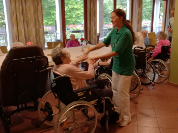 Eine lächelnde Pflegekraft tanzt mit einer älteren Person im Rollstuhl. Um sie herum sitzen weitere Bewohner:innen des Hauses. Im Hintergrund sind große Fenster mit Blick ins Grüne zu sehen. Die Szene vermittelt Lebensfreude und Herzlichkeit.