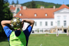 Eine Person im blauen Trikot und grünen Trainingsleibchen hält einen Fußball über dem Kopf zum Einwurf. Im Hintergrund sind eine Wiese, ein großes helles Gebäude mit rotem Dach, das Schloss Miller-Aichholz, und bewaldete Hügel zu sehen.
