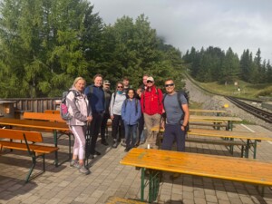 Eine Gruppe von neun wanderbereiten Personen steht auf einer gepflasterten Terrasse mit Holzbänken. Im Hintergrund sind Gleise einer Zahnradbahn, Bäume und leichter Nebel zu sehen. Die Gruppe lächelt in die Kamera.