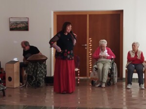 Im Vordergrund steht eine Frau mit Mikrofon, die ein Märchen erzählt. Links daneben sitzt ein Musiker und spielt ein rundes Instrument (Hang). Rechts im Bild sitzen mehrere ältere Personen, einige im Rollstuhl, und hören zu. Der Raum wirkt freundlich und ruhig.