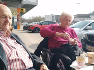 Ein älterer Bewohner sitzt an einem Tisch im Außenbereich eines Cafés, neben ihm sitzt eine Bewohnerin im Rollstuhl und isst Eis aus einem Becher. Auf dem Tisch stehen Eisbecher und Kaffeebecher, im Hintergrund sind parkende Autos zu sehen.