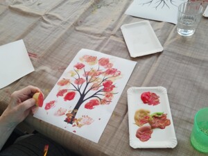 Auf dem Tisch liegt ein gemaltes Bild eines Baumes mit bunten Blättern. Eine ältere Person hält einen Pinsel und eine bemalte Kartoffel zum Stempeln. Daneben ein Farbteller mit herbstlichen Farben.