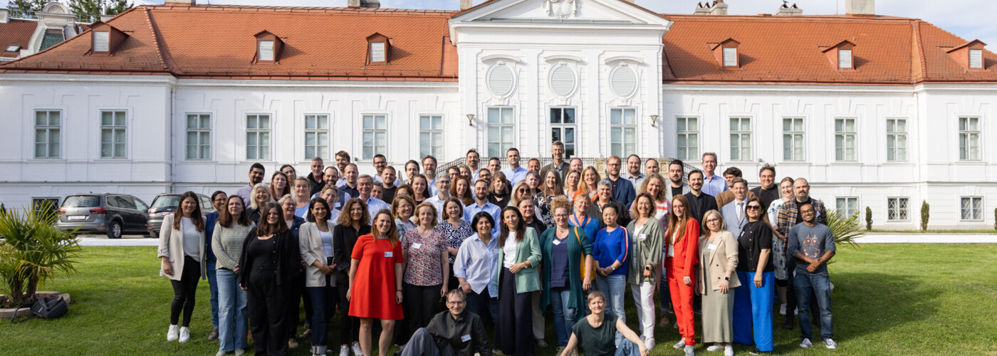 Über 80 Personen stehen auf einer Wiese vor einem weißen historischen Gebäude mit rotem Dach und posieren für ein gemeinsames Gruppenfoto.