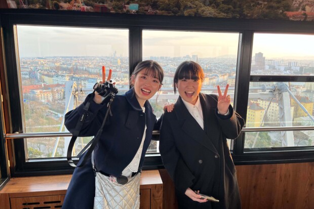 Zwei junge Japanerinnen stehen in einer verglasten Aussichtsgondel mit Blick über eine weitläufige Stadt bei warmem Abendlicht. Beide tragen dunkle Mäntel, lächeln in die Kamera und heben jeweils eine Hand zum Victory-Zeichen. Die linke Person hält zusätzlich eine Kamera, während im Hintergrund Dächer, Gebäude und Teile des Riesenrads bzw. der Gondelkonstruktion sichtbar sind.