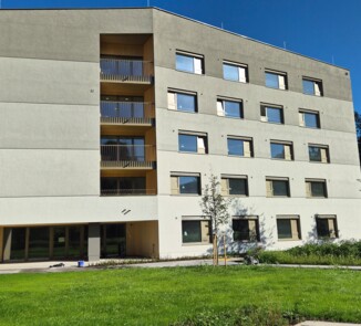 Ein fünfgeschossiges, modernes Wohngebäude mit vielen Fenstern, Balkonen und einer hellen Fassade. Es steht in einem gepflegten grünen Außenbereich mit Bäumen und Rasen.