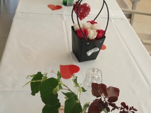 Ein langer, weiß gedeckter Tisch in einem hellen Gemeinschaftsraum ist mit roten Herzen, Blumensträußen, Gläsern und Wasserflaschen dekoriert. Die festliche Gestaltung bereitet die gemütliche Atmosphäre des Muttertagscafés für die Bewohner:innen vor.