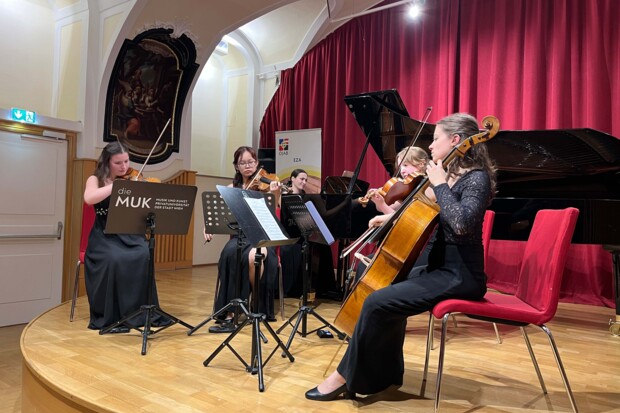 Vier Musiker:innen spielen gemeinsam Streichinstrumente in einem Ensemble. Sie sitzen auf Stühlen mit Notenständern vor sich. Im Hintergrund steht ein Flügel, und ein Schild mit der Aufschrift "die MUK" ist sichtbar.