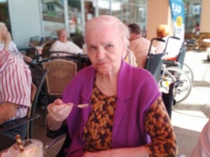 Eine Seniorin sitzt im Rollstuhl, hält einen Löffel mit Eis und schaut direkt in die Kamera. Vor ihr steht eine Glasschale mit Vanilleeis und einer Schokorolle.