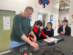 Eine Person erklärt an einem Arbeitstisch in einer Werkstatt den Umgang mit Glas als Werkstoff anhand kleiner Glaswürfel. Drei Teilnehmende stehen daneben und beobachten konzentriert die Demonstration. Auf dem Tisch liegen Werkzeuge wie Zangen, Kabel sowie eine Maschine, daneben befinden sich mehrere transparente Glasstücke. Die Umgebung ist technisch geprägt, mit Hinweisschildern, Geräten und einer funktionalen Einrichtung im Hintergrund.