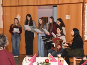 Sechs Jugendliche singen gemeinsam Weihnachtslieder, begleitet von Harmonika und Gitarre. Im Vordergrund sitzen ältere Personen und hören aufmerksam zu.