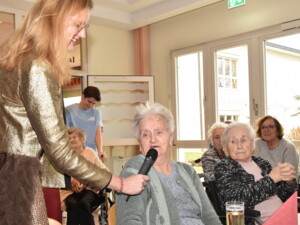 Eine ältere Bewohnerin trägt mit Mikrofon ein Weihnachtsgedicht vor. Eine Frau im goldenen Blazer reicht ihr das Mikrofon, im Hintergrund verfolgen andere Senior:innen und eine Pflegerin das Geschehen aufmerksam.