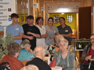 Pflegekräfte und Mitarbeiter:innen stehen singend und lachend in einer Türöffnung, während Bewohner:innen im Vordergrund an Tischen sitzen.