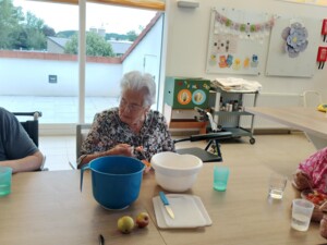 Mehrere Bewohner:innen sitzen an einem langen Tisch in einem hellen Aufenthaltsraum und essen Obstsalat aus kleinen Schüsseln. Auf dem Tisch stehen eine blaue und eine weiße Schüssel, Becher, ein Schneidebrett, ein Messer und einzelne Äpfel. Im Hintergrund sind große Fenster, eine Pinnwand, Bastelmaterialien und ein Rollator zu sehen.