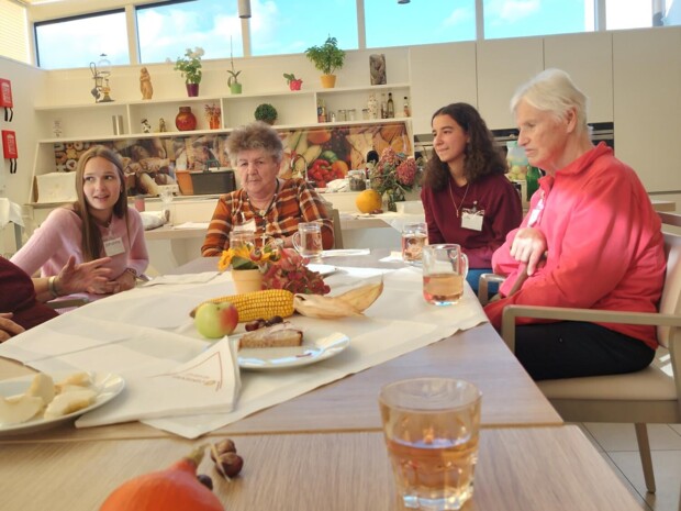 Junge Menschen und Senior:innen sitzen zusammen an einem Tisch, der herbstlich mit Obst, Mais und Getränken dekoriert ist. Im Hintergrund ist eine moderne Küche zu sehen.