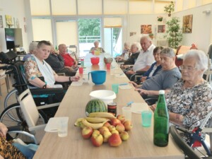Eine Gruppe von Bewohner:innen sitzt an einem langen Tisch mit Obst, Schüsseln und Bechern. Der helle Raum hat große Fenster, gelbe Rollos und mehrere Hilfsmittel wie Rollstühle und Rollatoren.