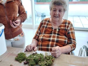 Eine ältere Person sitzt fröhlich an einem Tisch, auf dem Kastanien, Walnüsse, Moos und Rinde für eine Bastelarbeit verteilt sind. Neben ihr steht eine Betreuungsperson.