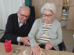 Ein älteres Paar arbeitet gemeinsam am Ausrollen von Teig. Vor ihnen liegen bereits ausgestochene Kekse in Blattform.