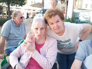 Eine Seniorin sitzt im Rollstuhl an einem Tisch mit Saftglas; eine Begleiter:in beugt sich lächelnd zu ihr. Weitere Marktbesucher:innen und ein "Kodak"-Schild im Hintergrund. Aufnahme vom August-Ausflug nach Güssing.