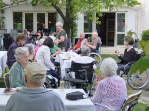 Viele Bewohner:innen und Angehörige sitzen im Garten an festlich gedeckten Tischen. Einige Personen nutzen Rollstühle, andere sitzen auf Stühlen. Im Hintergrund ist ein Musiker mit Akkordeon zu sehen, während die Gruppe in entspannter Atmosphäre beisammensitzt.