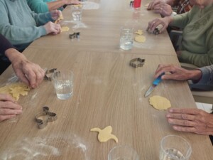 Zahlreiche Senior:innen sitzen an einem langen Tisch, bearbeiten Keksteig und verwenden verschiedene Ausstechformen.