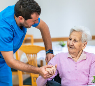Symbolbild. Ein junger Pflegefachkraft in blauer Berufskleidung hält die Hand einer älteren Person im Rollstuhl und spricht ihr ermutigend zu. Die Seniorin trägt einen rosa Pullover und lächelt ihm zu. Im Hintergrund sind Tische und Stühle in einem hellen Aufenthaltsraum zu sehen.