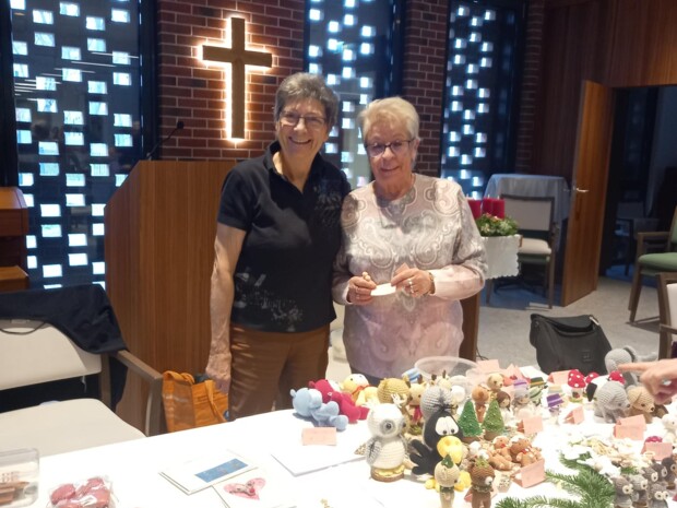 Zwei Bewohner:innen stehen hinter einem Tisch mit selbstgemachten Weihnachtsartikeln wie gehäkelten Figuren, Karten und Dekorationen. Im Hintergrund ist ein beleuchtetes Kreuz an der Wand zu sehen. Die beiden lächeln und präsentieren ihre Handarbeiten in einer festlich dekorierten Umgebung.