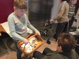 Ein Kind mit einem gefüllten Geschenkkorb überreicht einer älteren Person im Rollstuhl ein kleines Präsent. Im Hintergrund sind weitere Bewohner:innen mit Gehhilfen zu sehen.