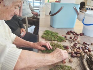Zwei ältere Menschen sitzen an einem Tisch, der mit Ästen, Moos, Rinde und weiteren Bastelmaterialien bedeckt ist. Eine Person ordnet Moos für eine Dekoration.