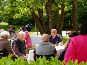 Bewohner:innen und Angehörige sitzen im grünen Garten an mehreren weiß gedeckten Tischen. Im Vordergrund rahmen helle Sträucher das Bild ein, darüber spenden große Bäume Schatten. Auf den Tischen stehen Getränke, Teller und kleine Frühlingsdekorationen.