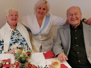 Zwei Bewohner:innen sitzen an einem Tisch. Eine Mitarbeiterin steht zwischen ihnen. Sie blicken in die Kamera und wirken entspannt. Die Tischdeko besteht aus Tannenzweigen und roten Elementen, was eine festliche Stimmung erzeugt.