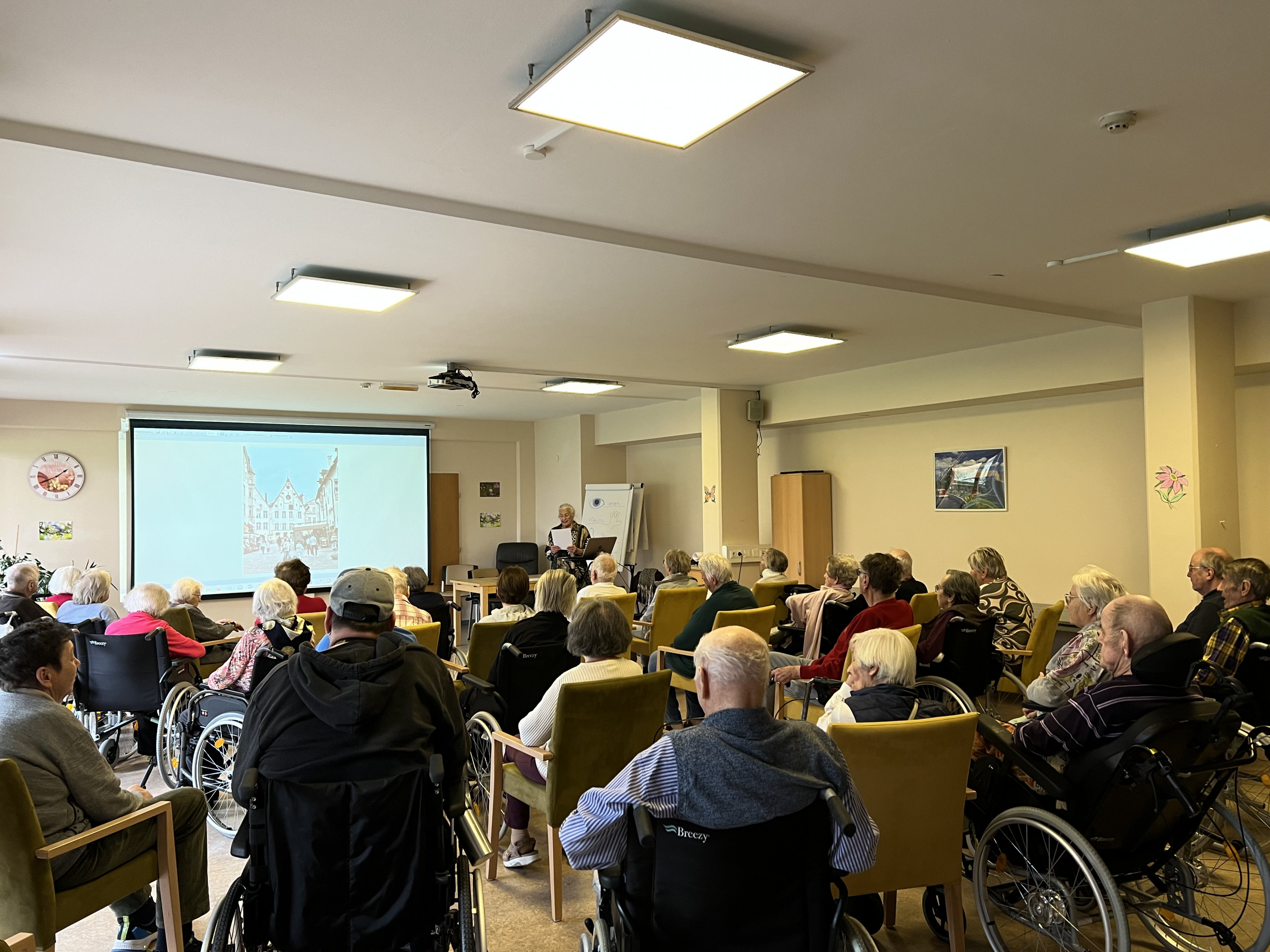 BewohnerInnen sitzend im Festsaal des Hauses beim Dia-Vortrag von Frau Klein, ganz hinten im Bild, stehend.