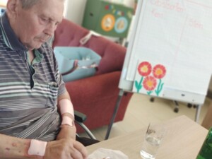 Ein älterer Bewohner sitzt am Tisch und bestreicht ein rechteckiges Stück Pizzateig mit Tomatensauce. Auf dem Tisch stehen ein Glas Wasser, ein Glas mit Tomatensauce und eine grüne Flasche. Im Hintergrund ist ein Flipchart mit einer Obstsortenliste zu sehen.