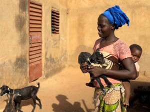 Eine erwachsene Frau trägt ein kleines Ziegenjunges im Arm, während ein Kleinkind auf dem Rücken getragen wird. Die Frau steht vor einem einfachen Gebäude mit Lehmwand und blickt lächelnd zur Seite. Im Vordergrund und Hintergrund sind weitere Ziegen zu sehen, die sich frei im Hof bewegen. Die Szene vermittelt den Alltag einer Familie mit Tierhaltung in einem ländlichen Umfeld.