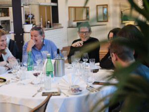 Mehrere Teilnehmende sitzen an einem gedeckten Tisch, lachen und unterhalten sich in entspannter Atmosphäre.