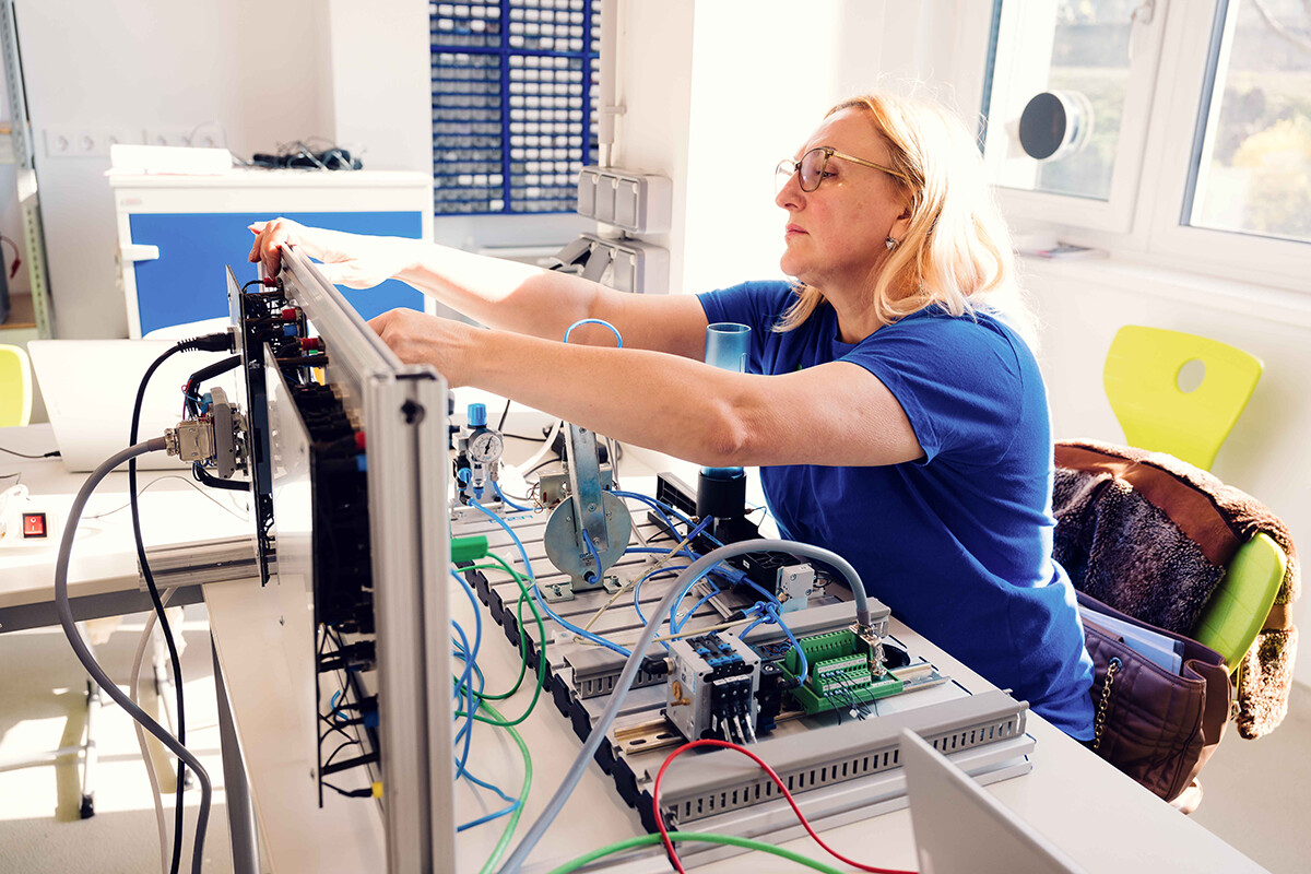 Eine Kursteilnehmerin in blauem Shirt sitzt an einem Arbeitstisch und justiert ein technisches Modell mit Kabeln, Schläuchen, Sensoren und Pneumatik-Bauteilen in einem hellen Schulungsraum. Foto: www.inframe.at