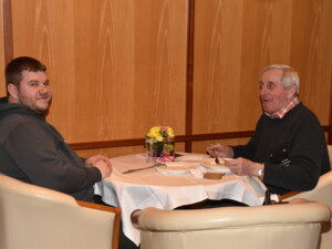 Zwei Männer unterschiedlichen Alters sitzen sich an einem kleinen runden Tisch gegenüber, essen und unterhalten sich. Der Tisch ist mit Blumen dekoriert.