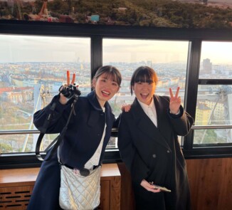 Zwei junge Japanerinnen stehen in einer verglasten Aussichtsgondel mit Blick über eine weitläufige Stadt bei warmem Abendlicht. Beide tragen dunkle Mäntel, lächeln in die Kamera und heben jeweils eine Hand zum Victory-Zeichen. Die linke Person hält zusätzlich eine Kamera, während im Hintergrund Dächer, Gebäude und Teile des Riesenrads bzw. der Gondelkonstruktion sichtbar sind.