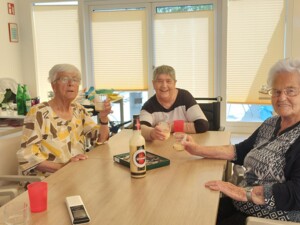 Mehrere Bewohner:innen sitzen gemeinsam an einem Tisch in der hellen "Kuchl". Einige halten Gläser in der Hand und lächeln in die Kamera. Auf dem Tisch stehen Getränke und Gläser; die Runde wirkt entspannt und freundlich.