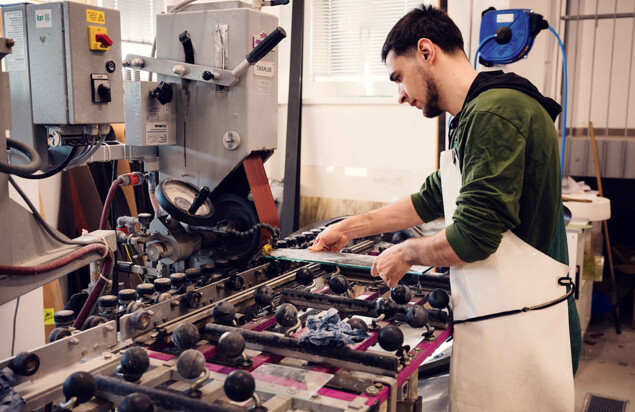 Ein Kursteilnehmer mit grünem Hoodie und weißer Schutzschürze führt eine Glasplatte vorsichtig durch eine große Maschine mit vielen Rollen und Bedienelementen. Foto: www.inframe.at