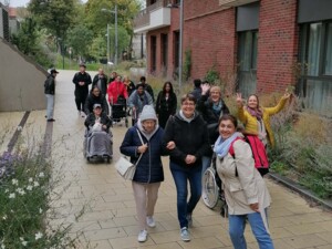 Eine Gruppe von Bewohner:innen und Begleiter:innen geht gemeinsam auf einem Gehweg entlang eines Gebäudes. Einige Personen schieben Rollstühle, andere gehen zu Fuß. Mehrere Teilnehmende lächeln und winken in die Kamera. Die Umgebung zeigt Bäume, Wohnhäuser und eine herbstliche Stimmung.