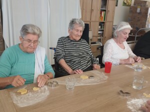 Drei Senior:innen sitzen an einem langen Tisch und bereiten Kekse vor. Auf dem Tisch liegen Mehl, Ausstechformen und Teigstücke.
