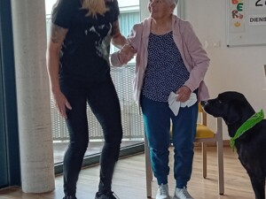 Eine Betreuer:in hält die Hand einer älteren Person beim Gehen. Beide lächeln sich an. Therapiehund Buddy mit grünem Halstuch steht daneben und beobachtet die Szene.