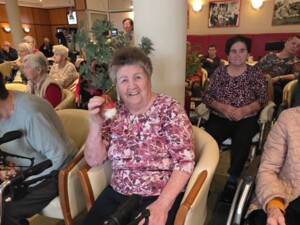 Eine ältere Frau hält einen weihnachtlichen Anhänger in der Hand und lächelt in die Kamera. Um sie herum sitzen weitere Bewohner:innen mit Rollatoren und Rollstühlen.
