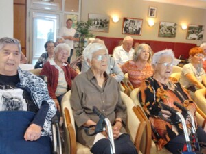 Eine Gruppe von Bewohner:innen sitzt in Stühlen und Rollstühlen in einem Gemeinschaftsraum. Einige schauen konzentriert nach vorne, andere wirken, als würden sie mitsingen.