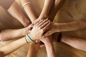 Mehrere Arme und Hände von Personen, die übereinandergelegt in einem Kreis auf einer Holzoberfläche ruhen - ein Symbol für Teamarbeit, Zusammenhalt und Diversität.