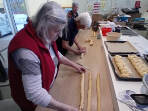 Drei Senior:innen arbeiten gemeinsam an einem Tisch, rollen Teigstränge und bereiten diese zum Flechten der Striezel vor. Im Hintergrund sind vorbereitete Teiglinge sichtbar.