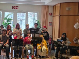 Schüler:innen spielen verschiedene Musikinstrumente wie Trompeten, Klarinette, Gitarre und Keyboard. Andere singen oder lesen Weihnachtsgeschichten vor. Alle tragen festliche oder alltägliche Kleidung, manche mit Weihnachtsmützen.
